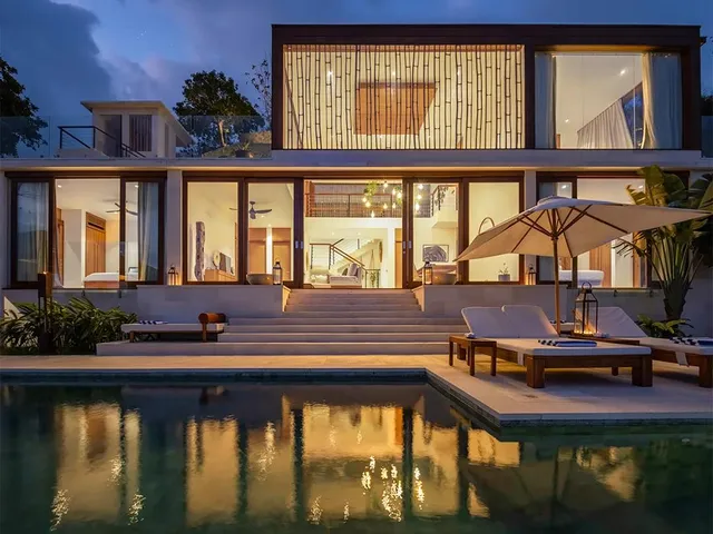 A two-storey Selo villa lit at night, its glass walls reflected in the pool in front, with loungers under a parasol.