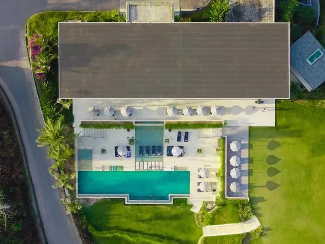 An aerial view of a resort: a long roof over a pool deck with parasols and loungers, lawns and palms around it.