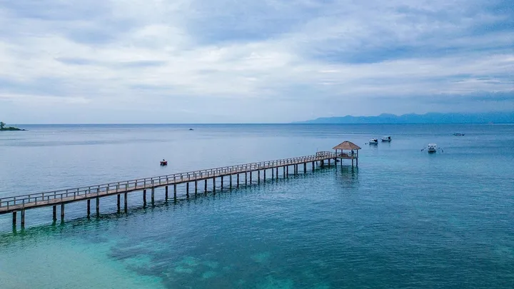 Aerial view of a long wooden jetty on stilts running over clear turquoise shallows to a thatched shelter, with small boats moored beyond it under a grey sky.